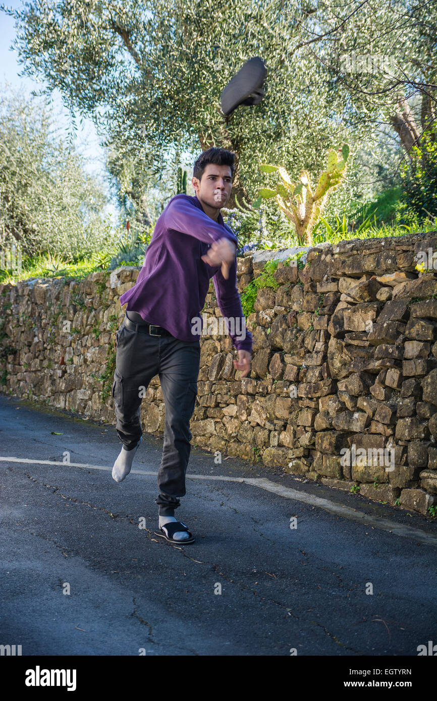 Jeune homme en colère, debout au milieu d'une route rurale avec un mur de pierre de lancer sa chaussure grâce à l'air vers l'appareil photo Banque D'Images