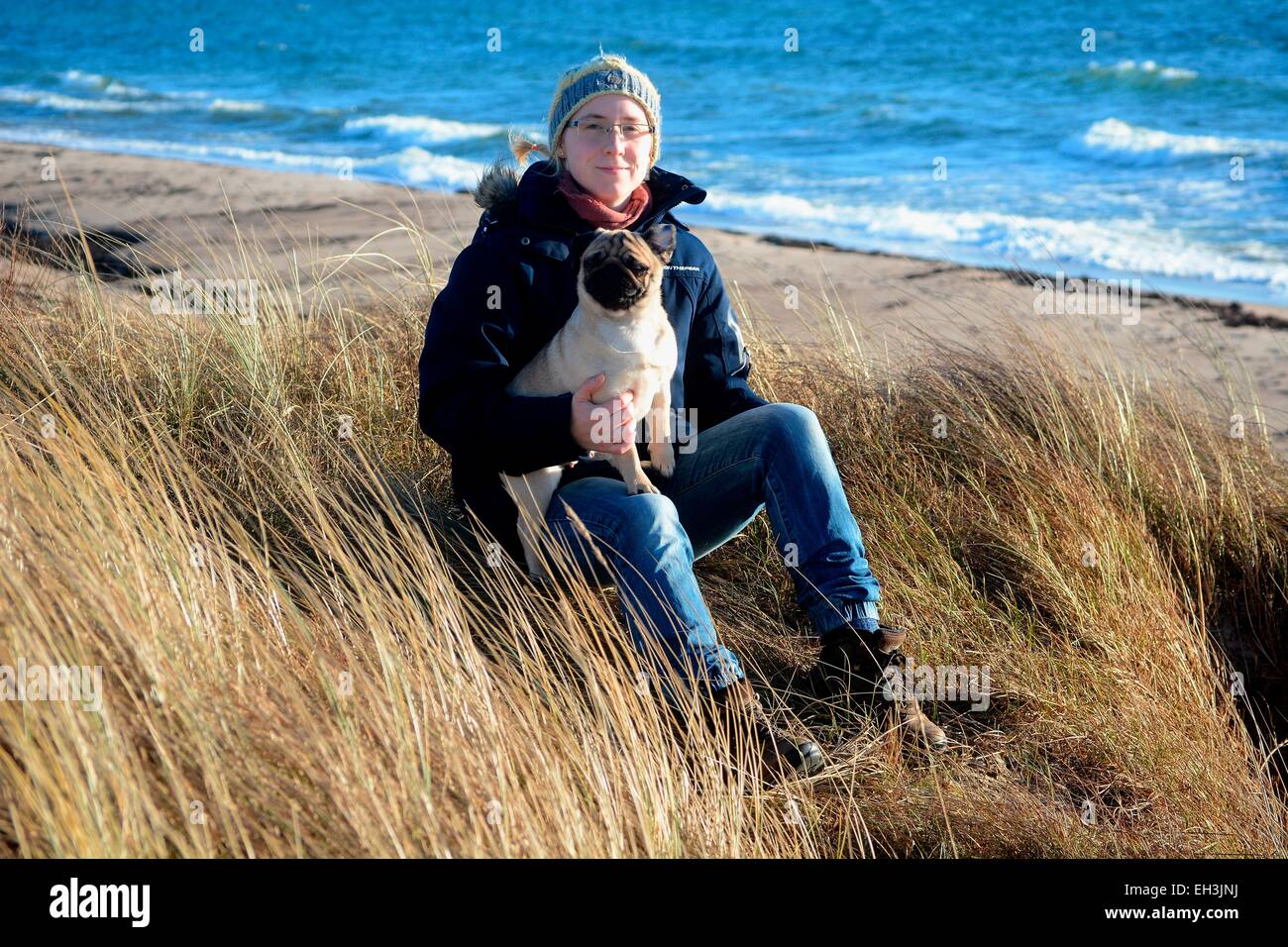 Jeune femme portant des vêtements d'hiver et la tenue d'un pug, assis dans l'herbe sèche à la mer Baltique, Scania, Suède Banque D'Images