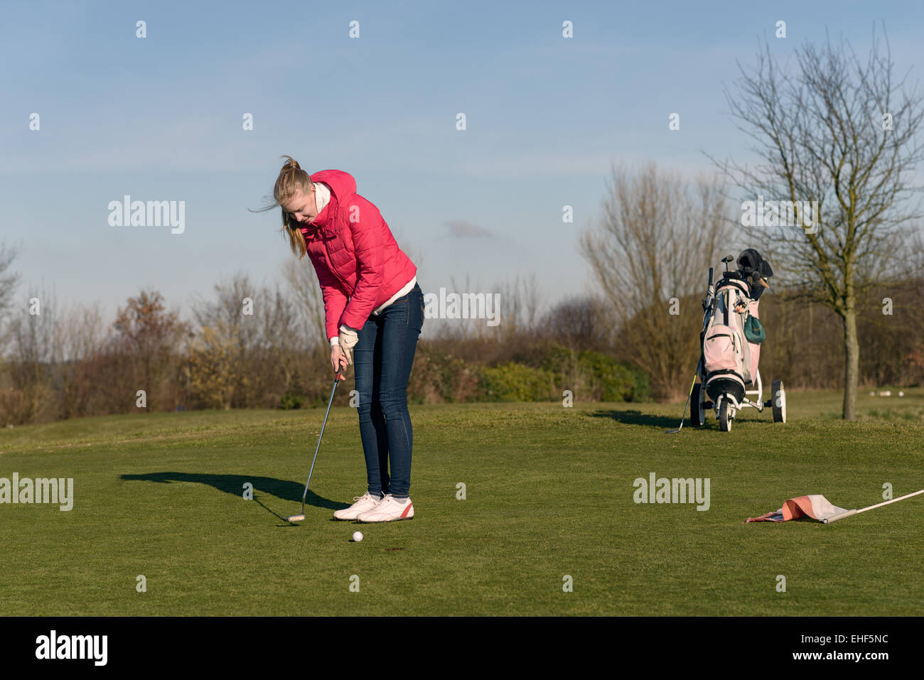 Woman alignement d'un putt sur le green avec son chariot de golf et clubs visible derrière sur un ciel bleu ensoleillé jour Banque D'Images