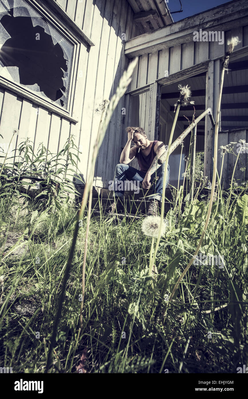 Un jeune homme assis à l'extérieur d'une maison abandonnée et la fumée de cigarettes. Banque D'Images