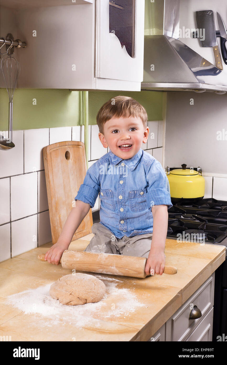 Beau garçon assis sur le comptoir de la cuisine tenant un rouleau à pâtisserie. faire du pain ou des pizzas. Banque D'Images