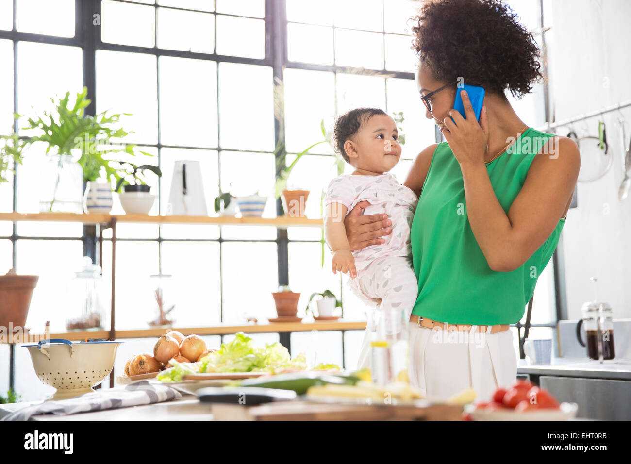 Mère tenant son bébé fille, alors qu'à l'aide de mobile phone in kitchen Banque D'Images