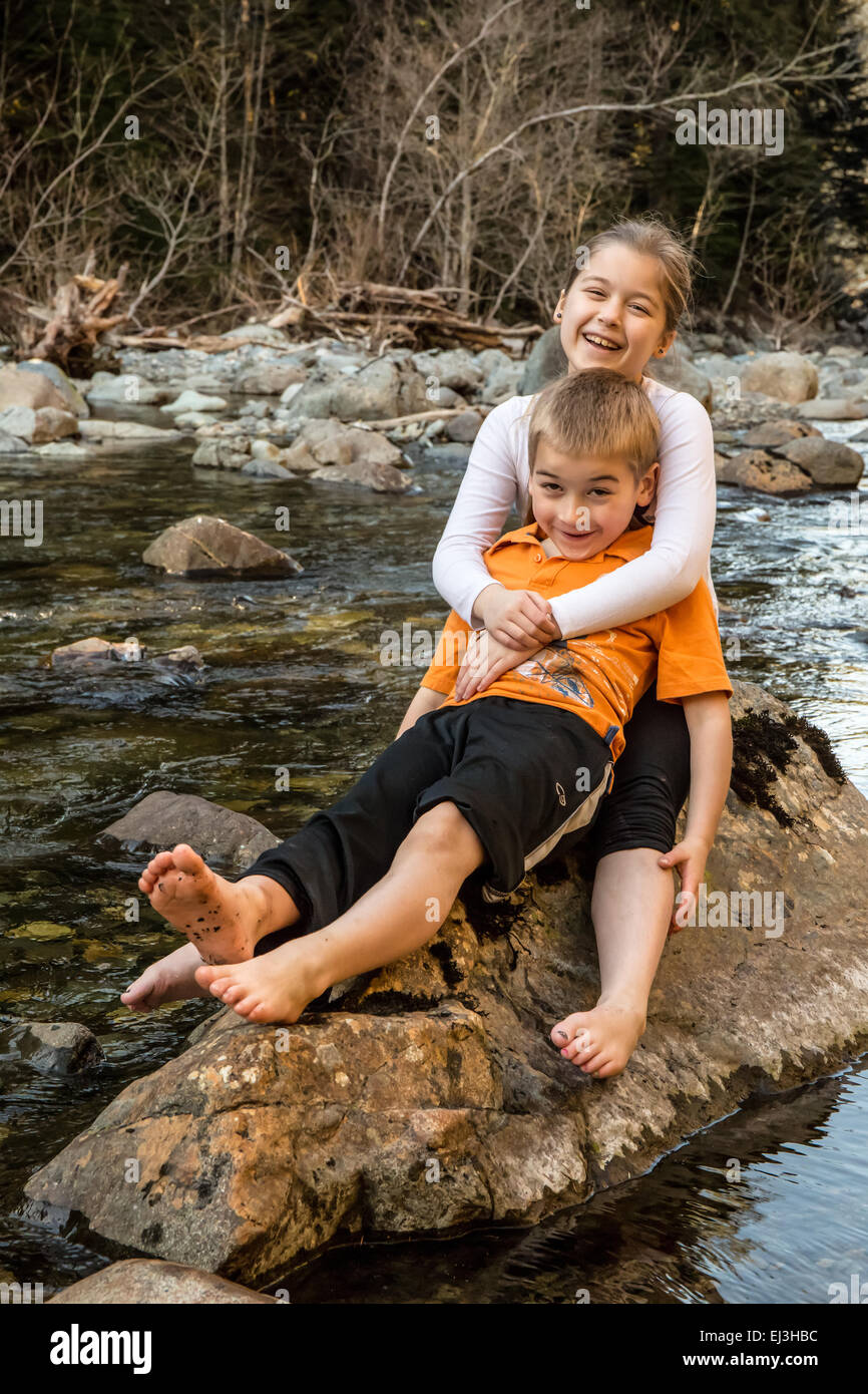 Neuf ans, fille de donner son frère de sept ans une accolade, assis sur un rocher dans une rivière peu profonde Banque D'Images