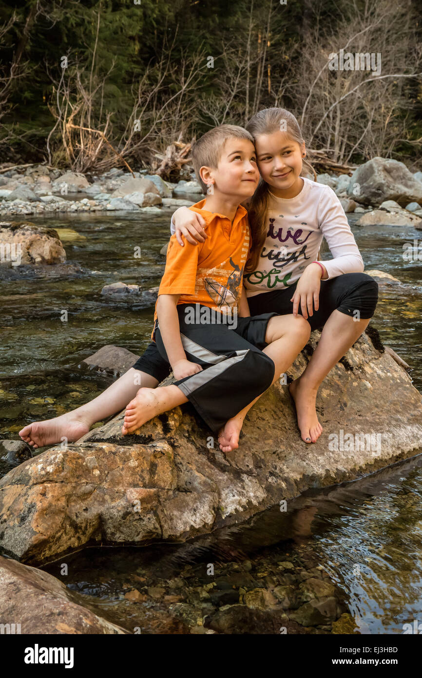 Neuf ans, fille de donner son frère de sept ans une accolade, assis sur un rocher dans une rivière peu profonde Banque D'Images