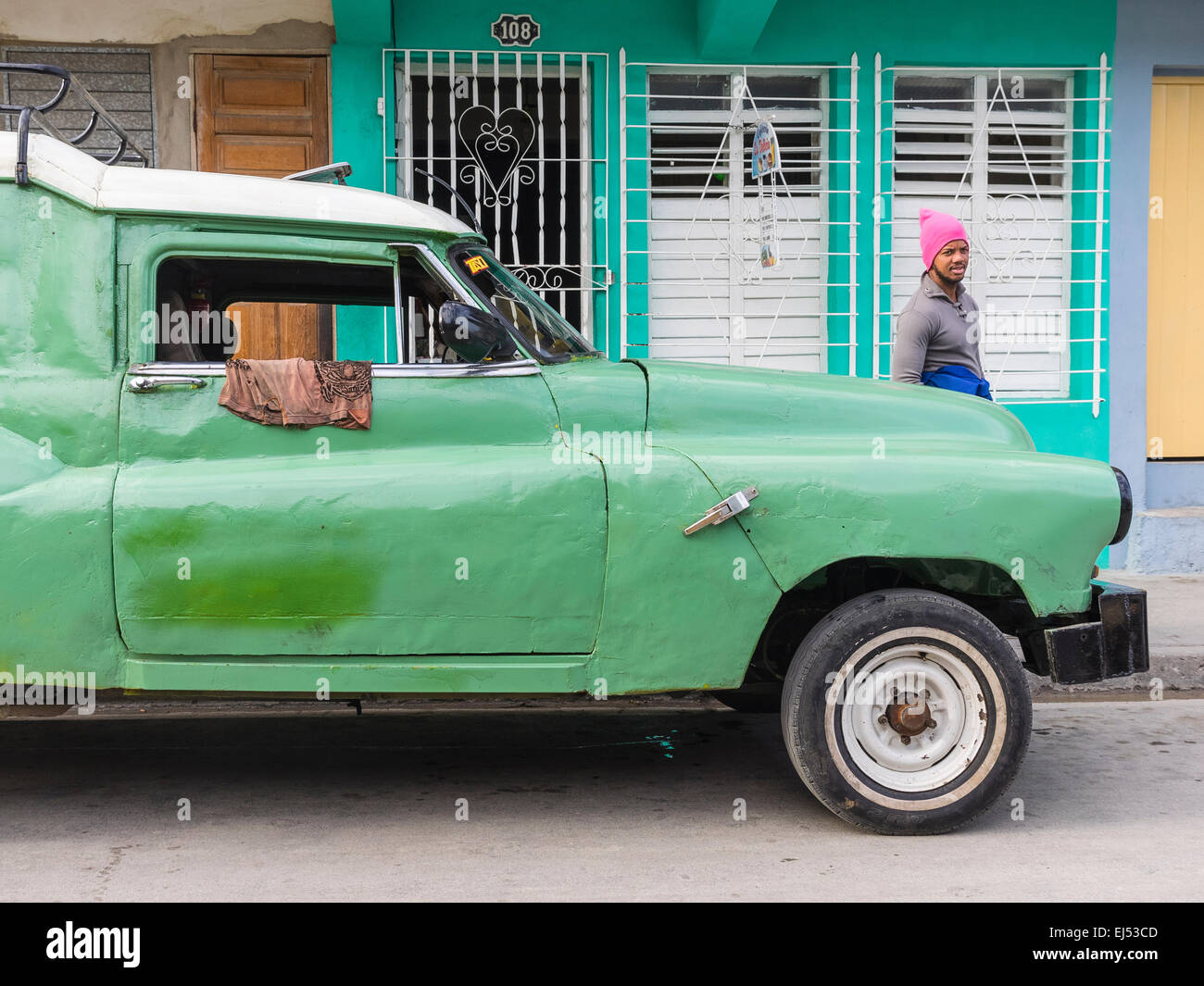 Afro-cubain un homme portant un chapeau en laine rose chaud s'en tient à un livre vert et blanc beat-up vieille voiture à Santiago de Cuba. Banque D'Images