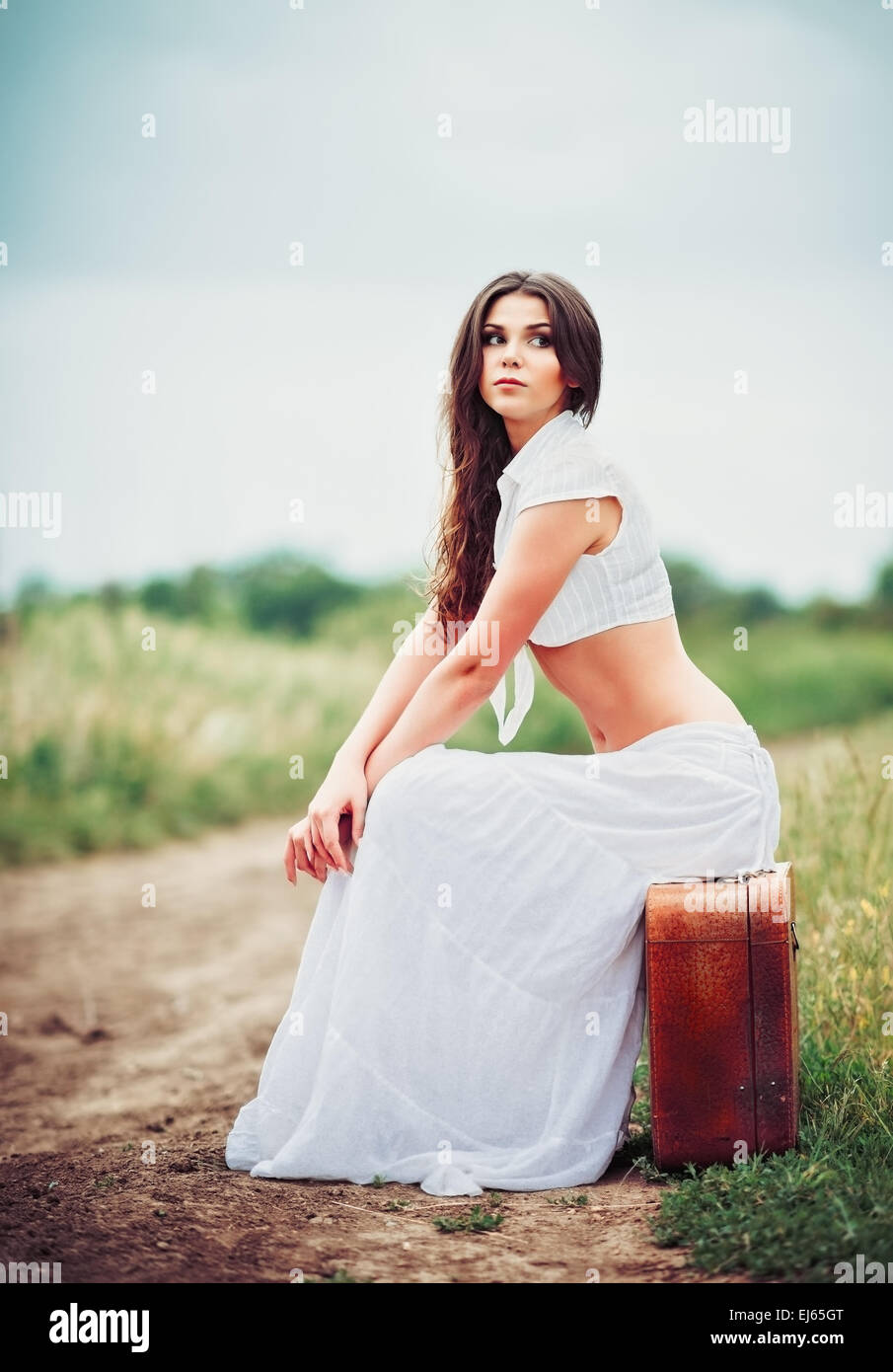 Belle jeune femme est assise sur une valise près de field road Banque D'Images