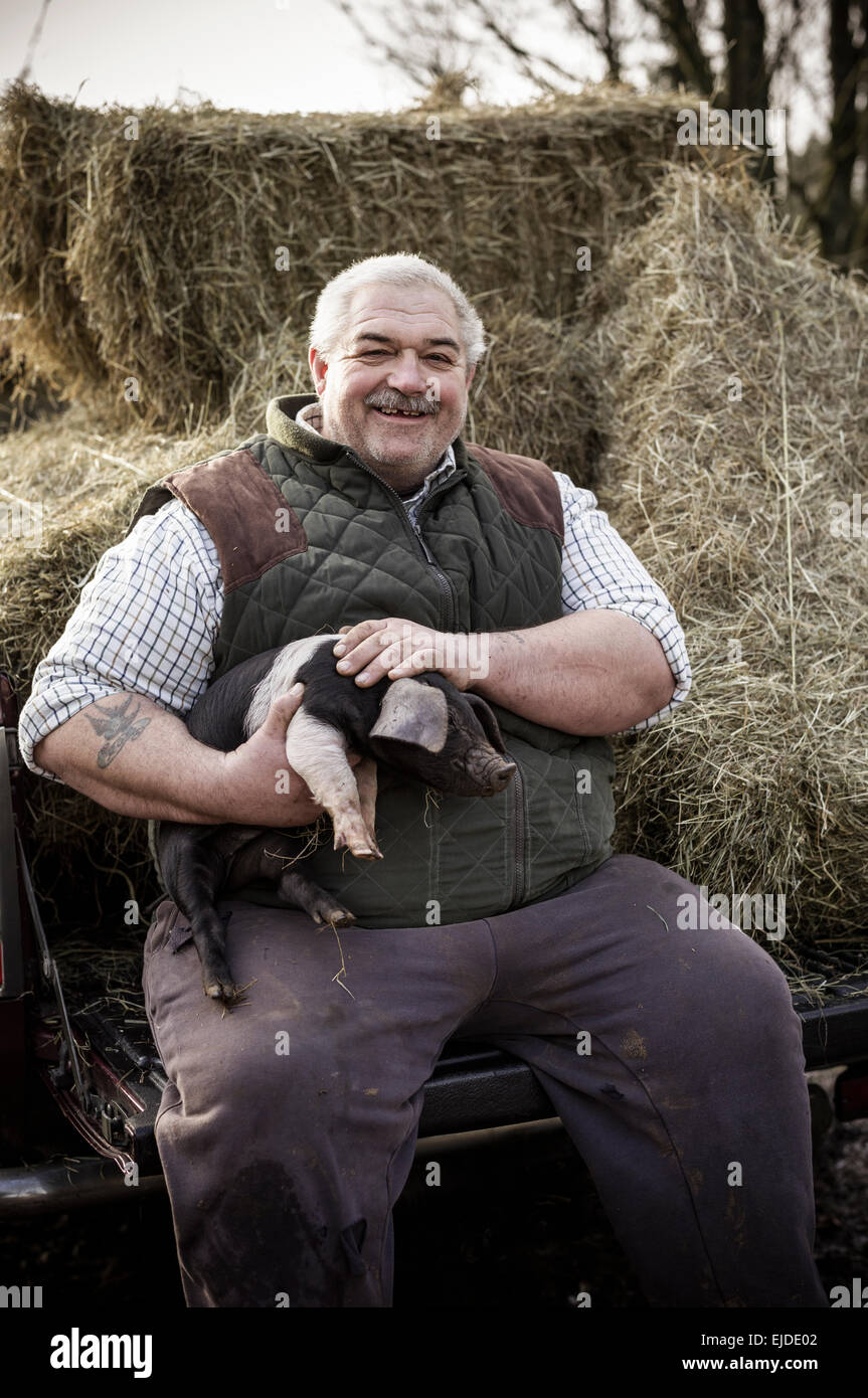 Un grand homme, agriculteur dans un gilet et vêtements de travail, tenant un porcelet et souriant Banque D'Images