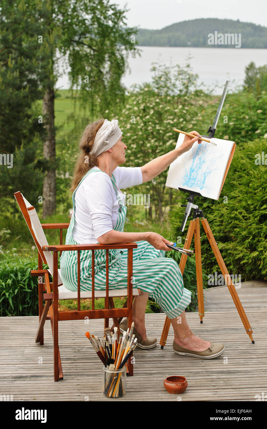Senior woman painting à l'extérieur. Elle siège dans une toile chaise devant le chevalet. La journée est couvert en donnant une lumière douce Banque D'Images