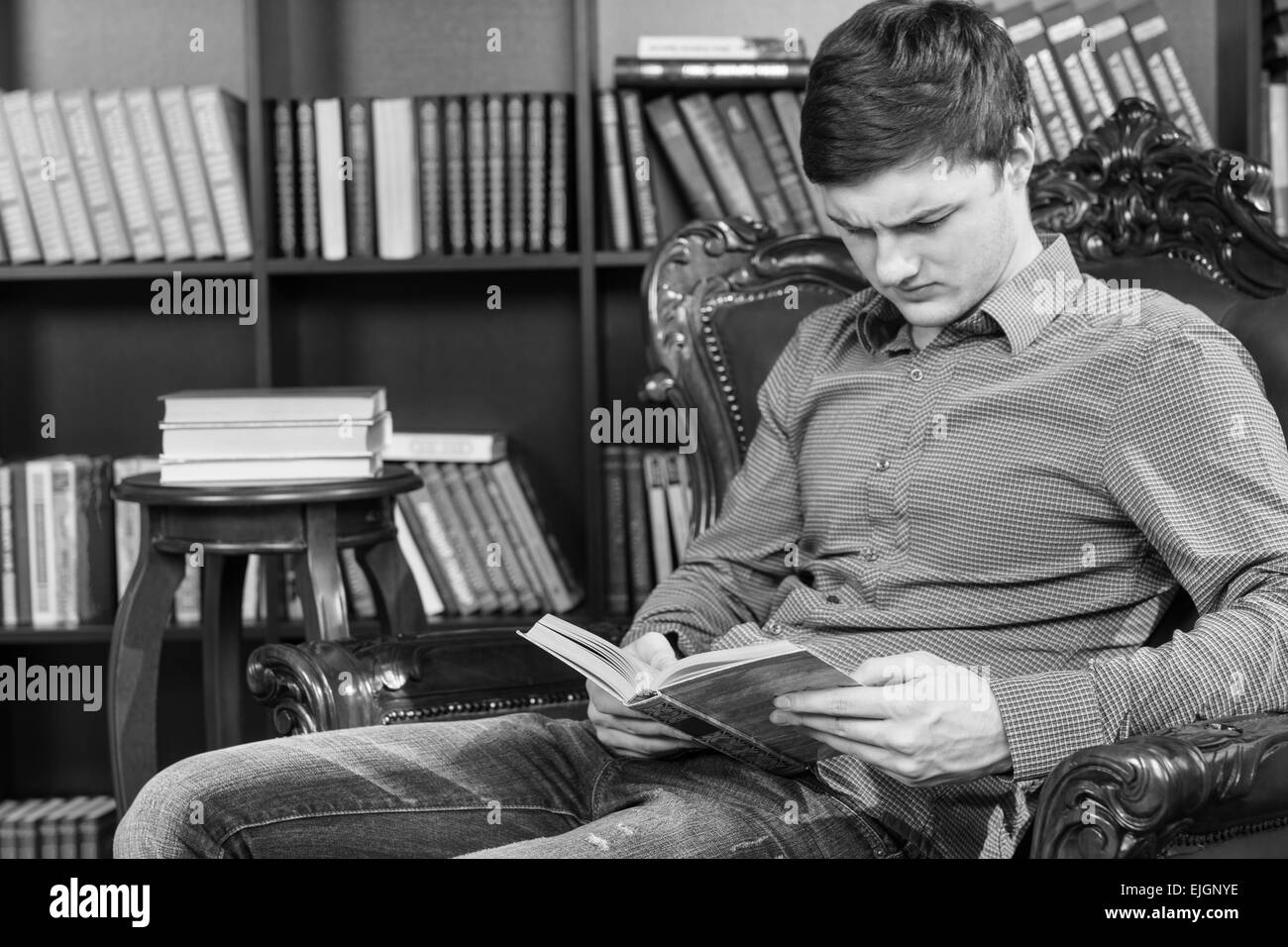 De graves beau jeune homme assis sur une chaise près de la bibliothèque tout en lisant un livre au sérieux, capturé en monochrome. Banque D'Images