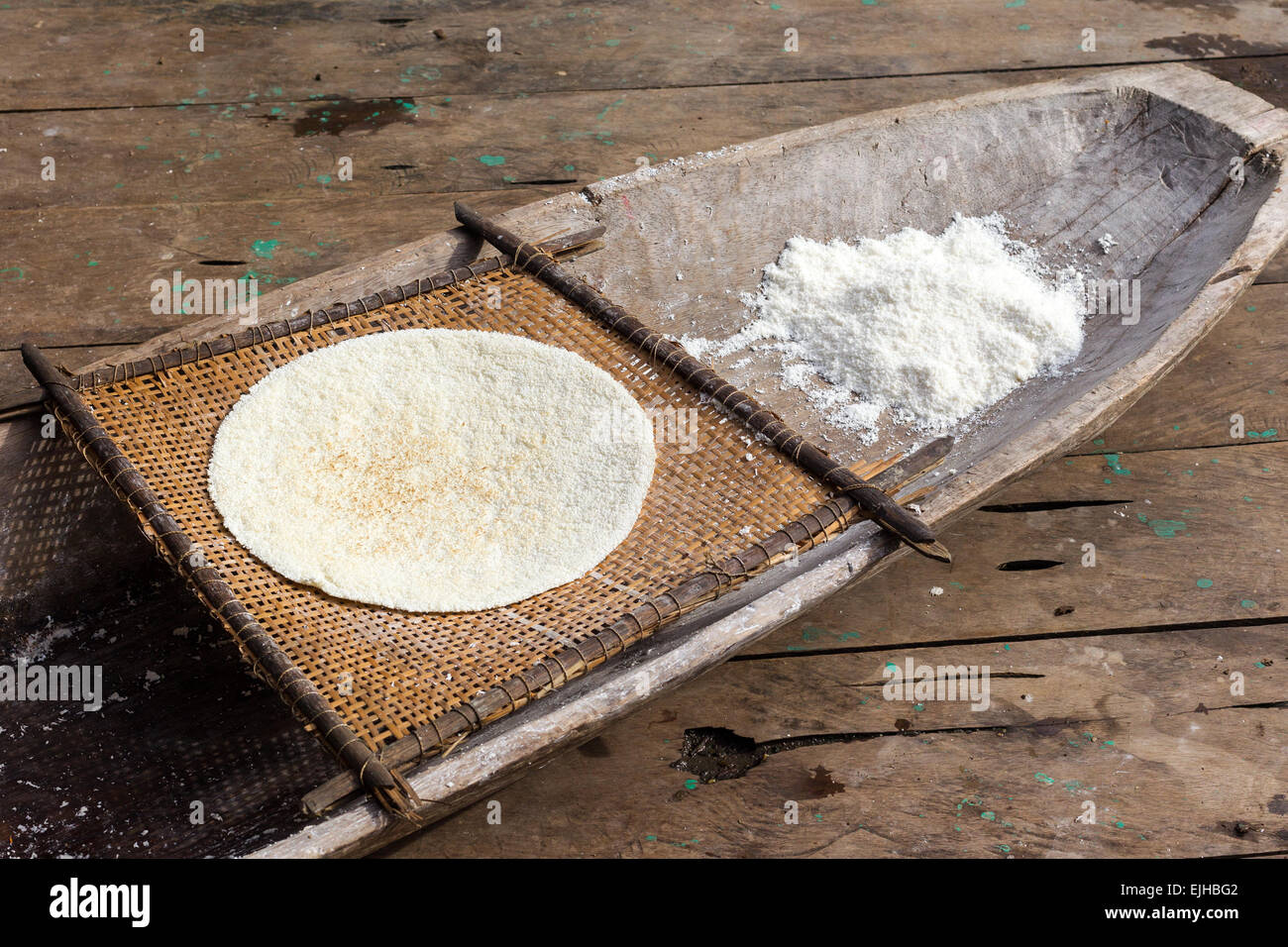 Un disque circulaire et plat de farine de riz blanc repose sur un tapis de bambou tissé, qui est placé à l'intérieur d'un plateau en bois altéré. De la farine de riz en vrac supplémentaire est empilée dans le plateau à côté du tapis. Le tout Banque D'Images