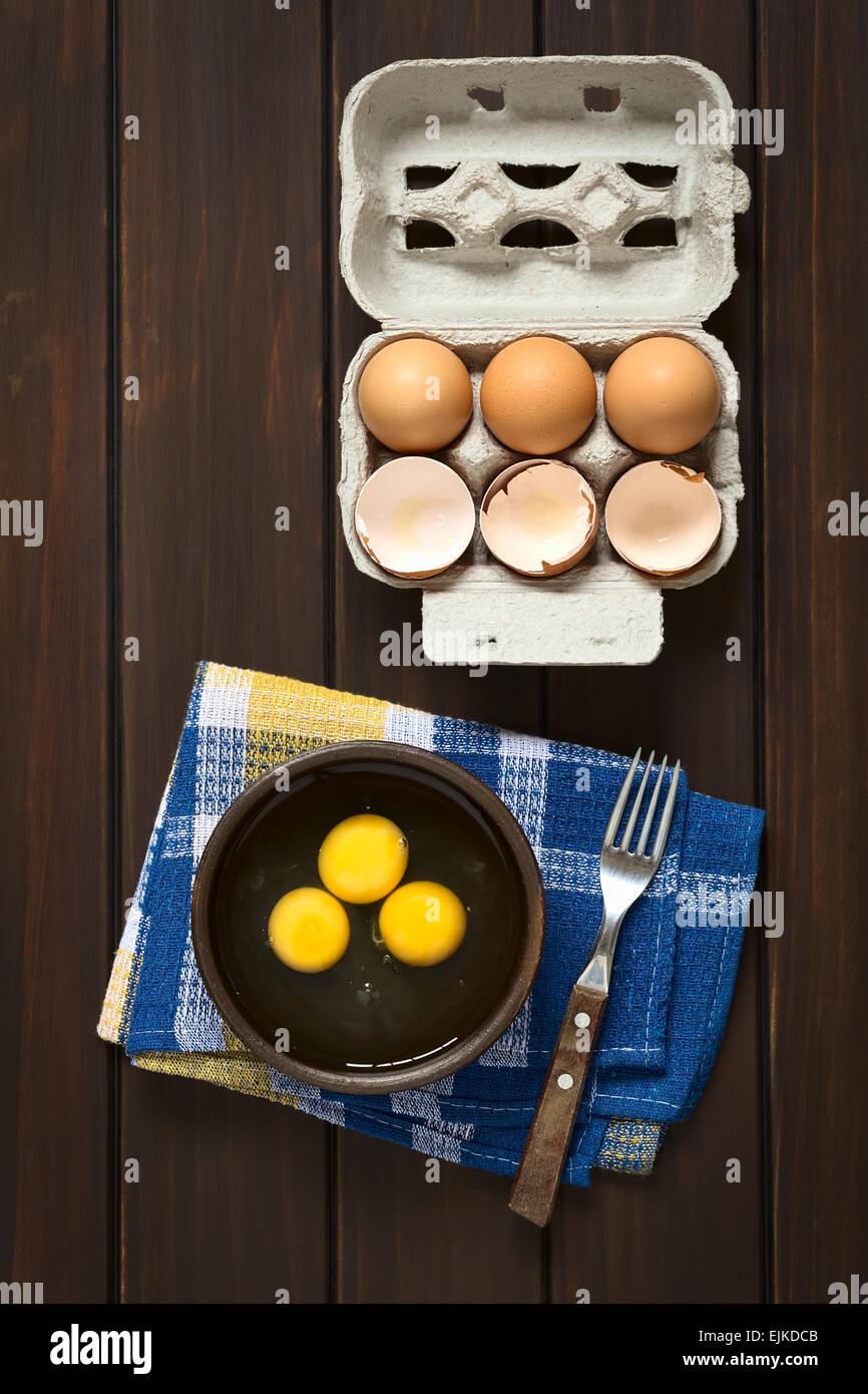 Coup de frais généraux trois oeufs crus dans bol rustique avec une fourchette sur le côté et des oeufs et des coquilles d'oeufs avec la boîte au-dessus Banque D'Images