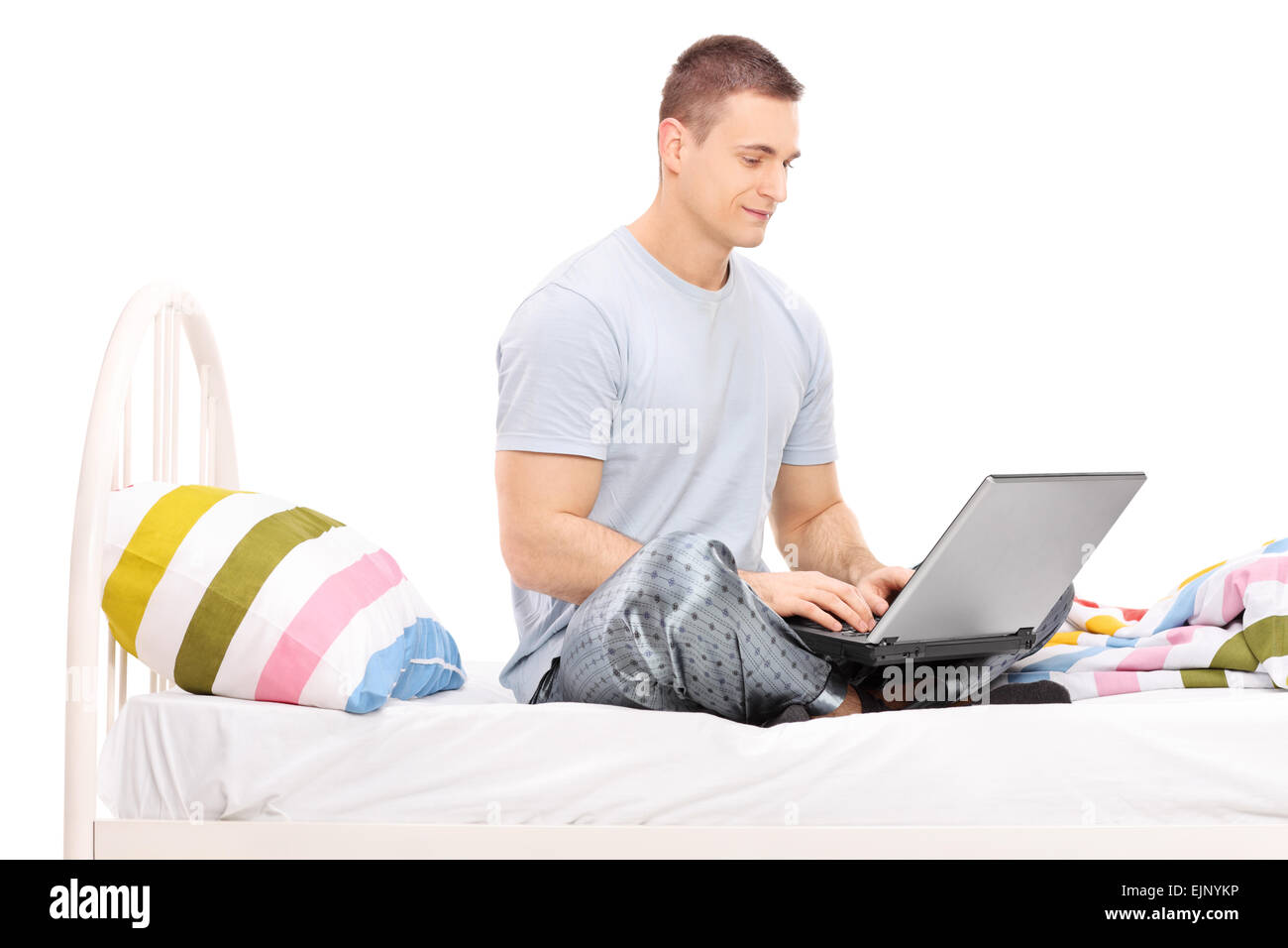 Portrait d'un jeune homme en pyjama de travailler sur un ordinateur portable assis sur un lit isolé sur fond blanc Banque D'Images Portrait d'un jeune homme en pyjama de travailler sur un ordinateur portable assis sur un lit isolé sur fond blanc Banque D'Images