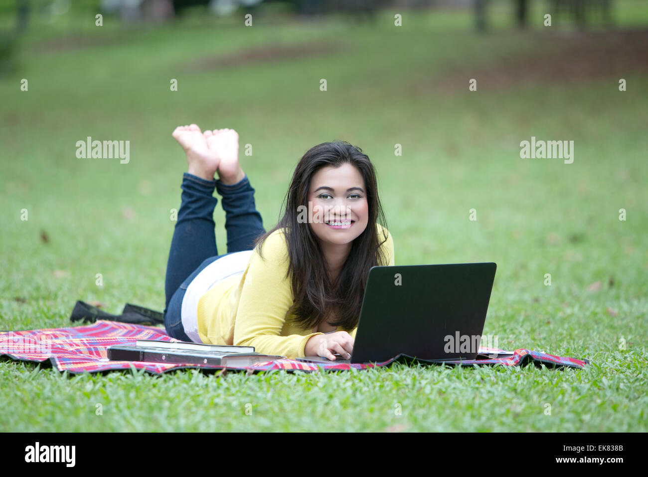 Belle jeune femme d'origine asiatique ou de l'université college student lying on stomach avec des livres, des dossiers et un ordinateur portable dans un parc. Banque D'Images