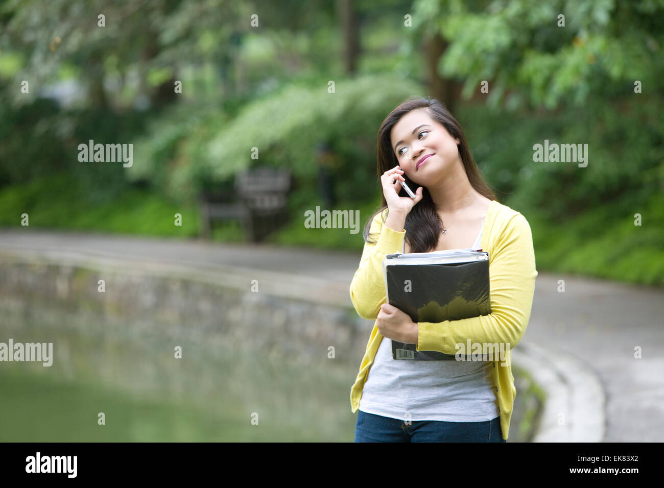Belle jeune femme à l'université avec des livres et des fichiers dans le bras, en utilisant son téléphone, debout dans un parc. Banque D'Images