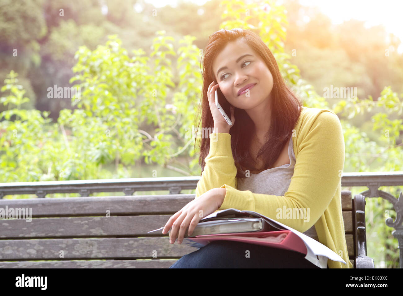 Belle jeune femme à l'université avec des livres et des dossiers sur les genoux, en utilisant son téléphone, assis sur le banc en bois dans un parc. Banque D'Images