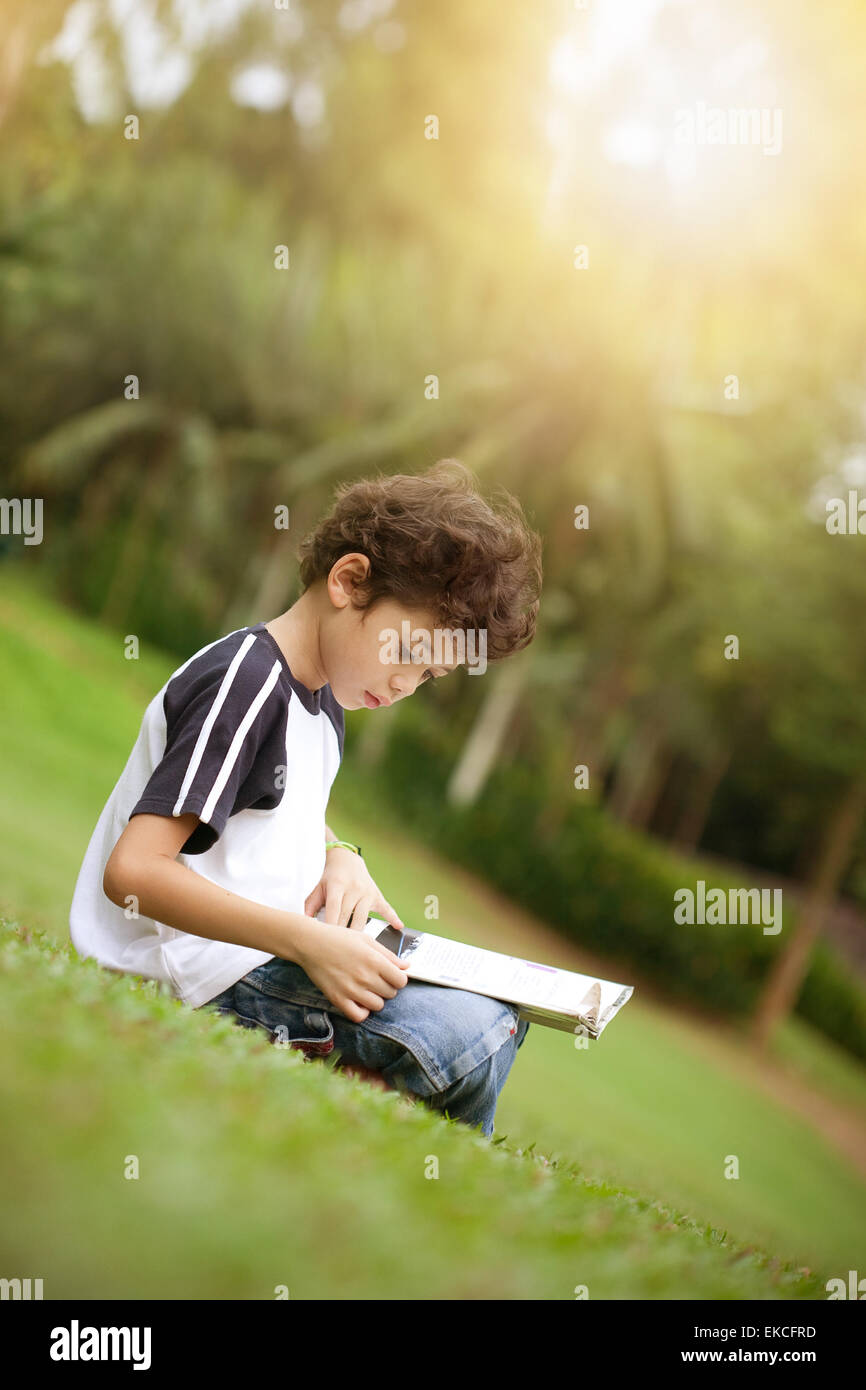 Jeune garçon pan asiatique apprécie sa lecture réserve en parc extérieur au soleil Banque D'Images
