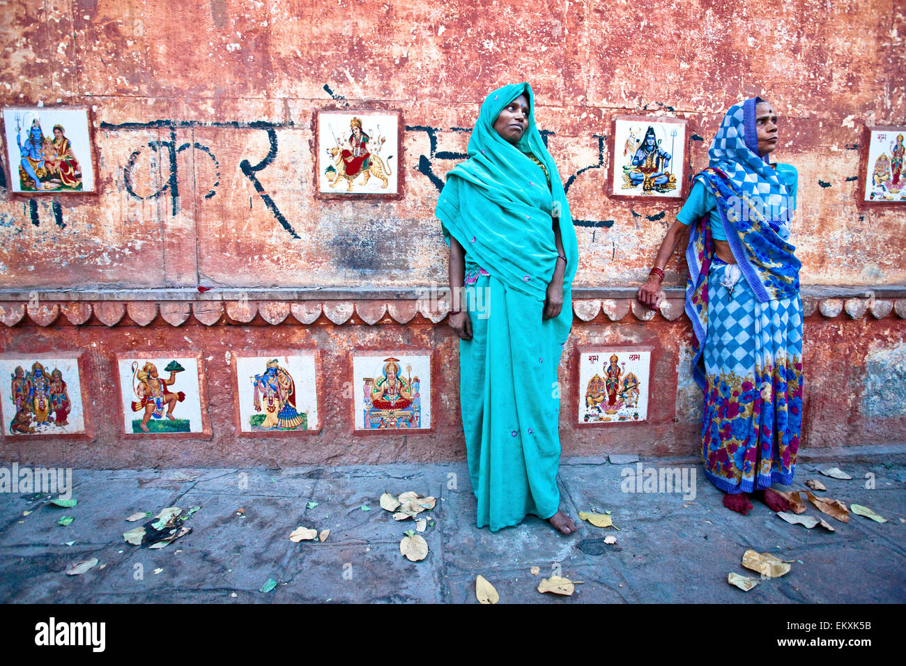 Deux femmes en saris debout dans la rue contre un mur bordé d'images de dieux hindous ; Varanasi Inde Banque D'Images