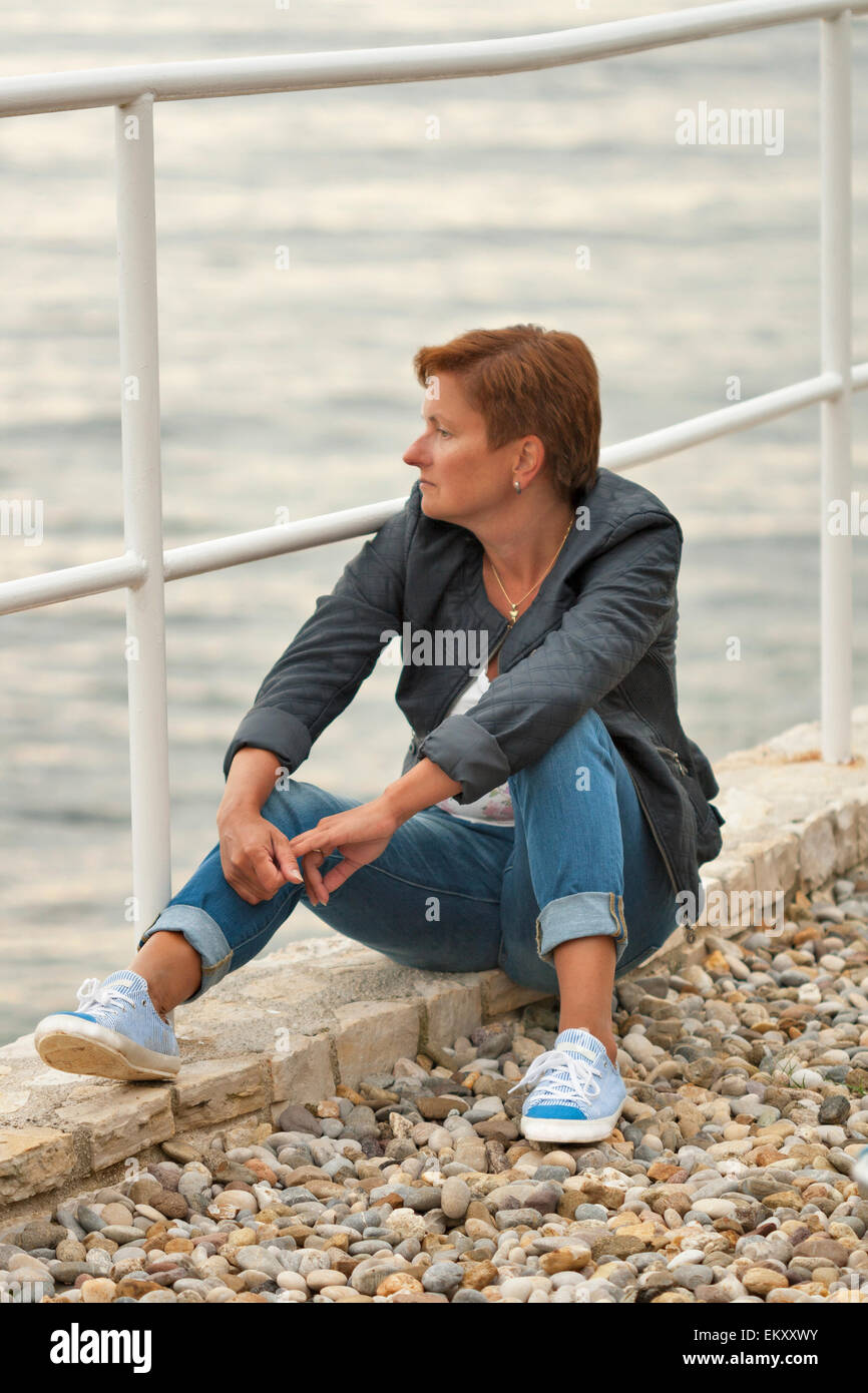 Middle aged woman femme bronzée en automne vêtements assis sur la plage de la mer de rochers Banque D'Images