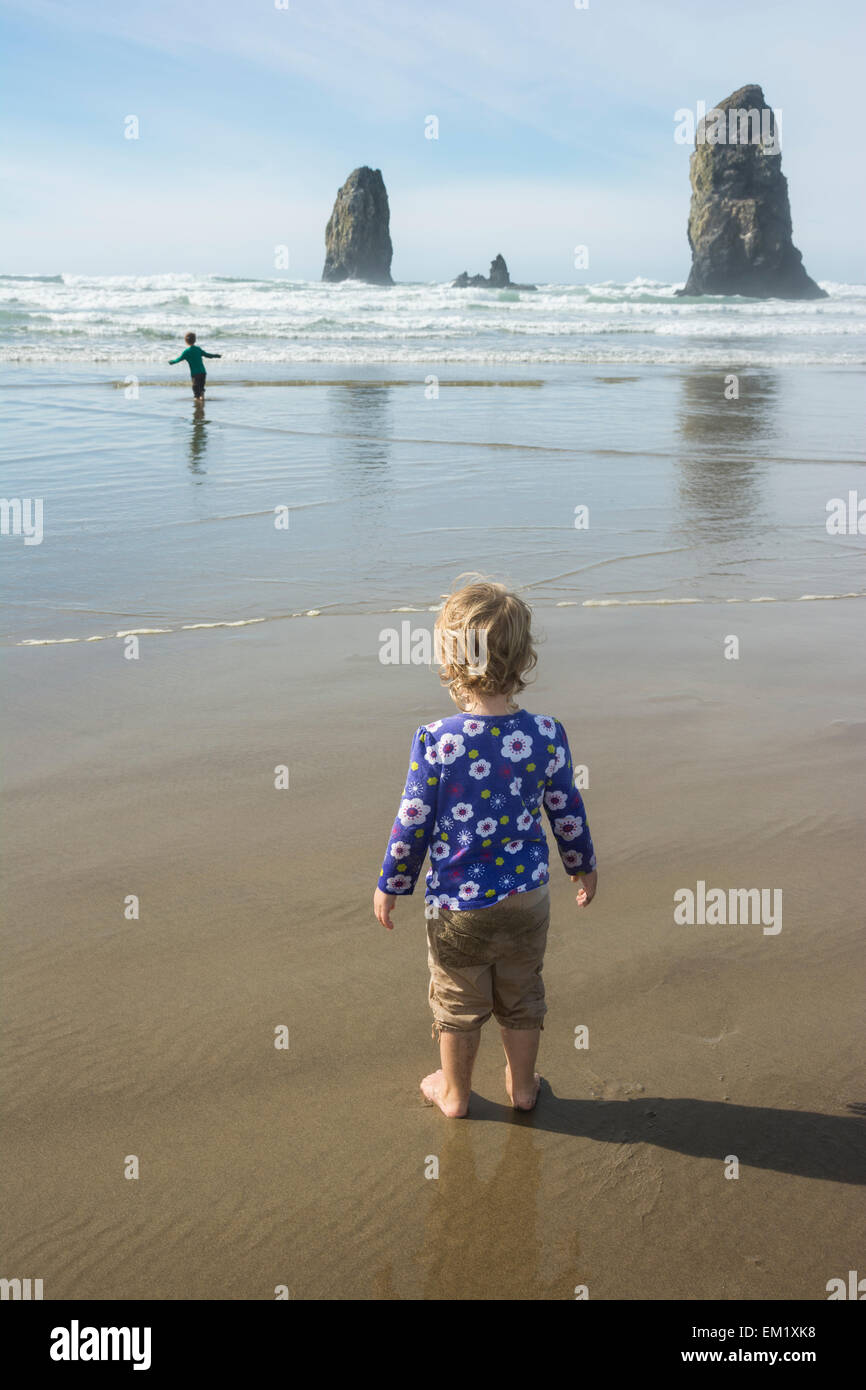 À partir de la rive, bébé fille voit son frère tester les vagues de l'océan froide, Cannon Beach, Oregon. Banque D'Images