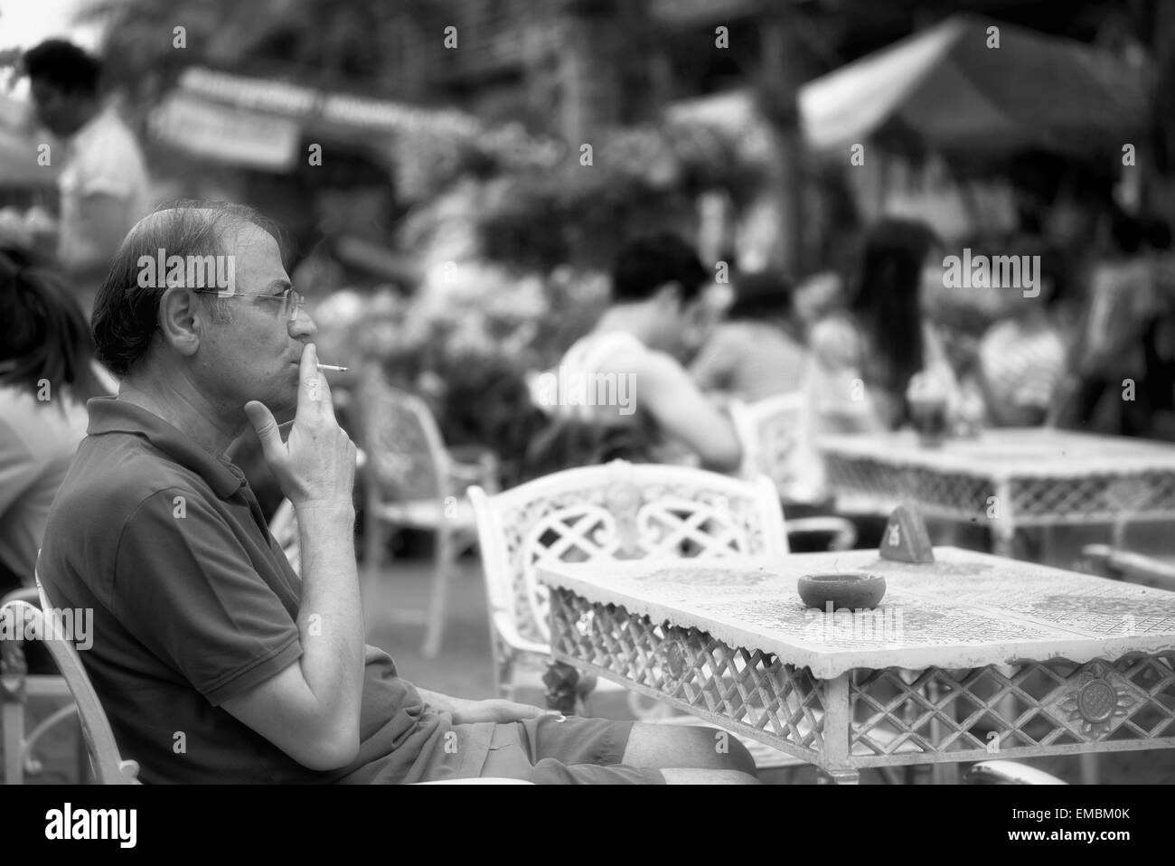 L'homme fume une cigarette et assis seul à une table de café en bordure de route. Photographie noir et blanc Banque D'Images