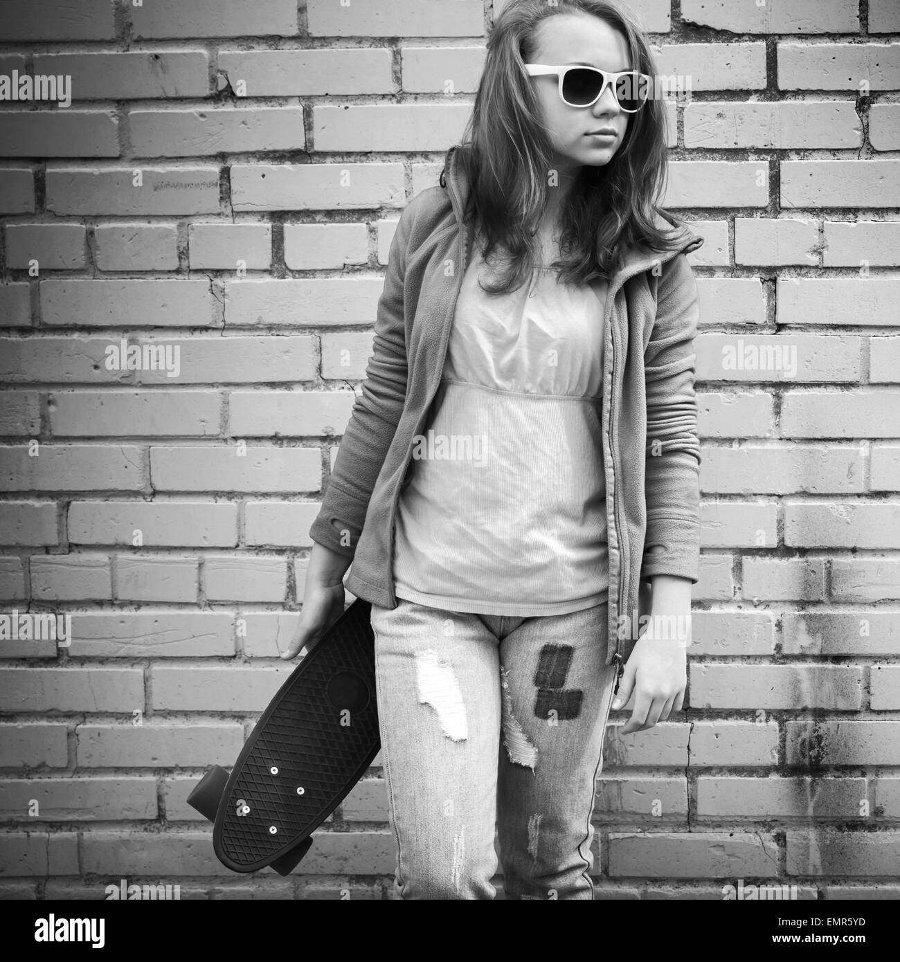 Adolescente blonde en jeans et lunettes détient plus de skateboard urban mur de briques, l'arrière-plan photo monochrome Banque D'Images