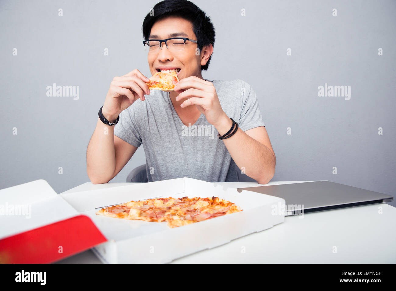 Young Asian man eating pizza au bureau au fond gris Banque D'Images