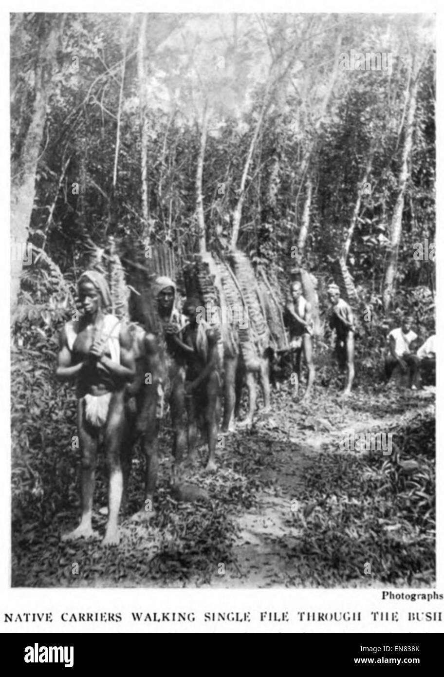 Une photographie de 1922 représentant des transporteurs autochtones marchant une seule file dans la brousse, capturant les méthodes de transport traditionnelles dans l'Afrique coloniale. Banque D'Images