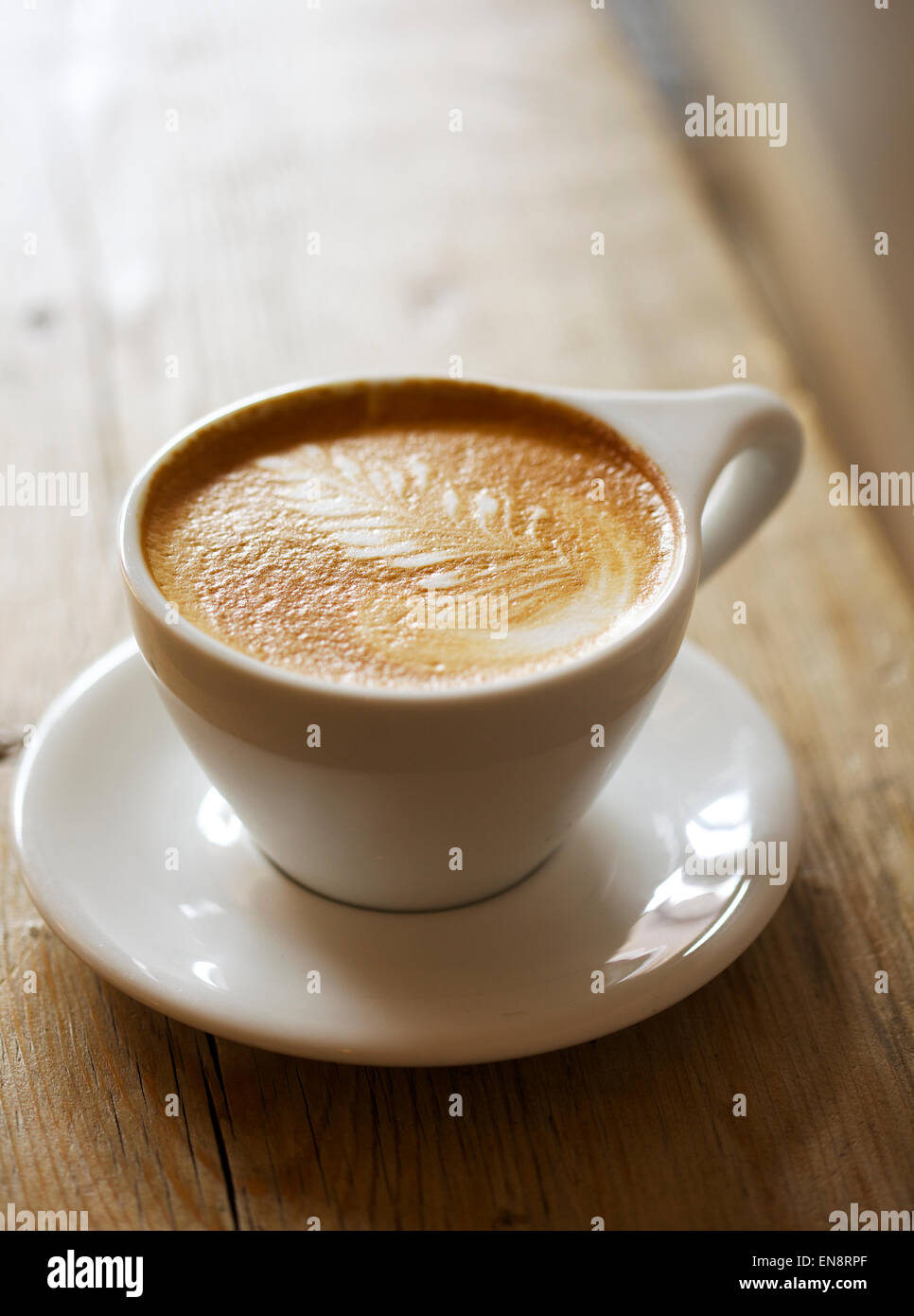 Un cappuccino avec un motif de feuilles dans la mousse dans une tasse blanche avec une soucoupe assis sur une table en bois, l'arrière-plan est flou. Banque D'Images