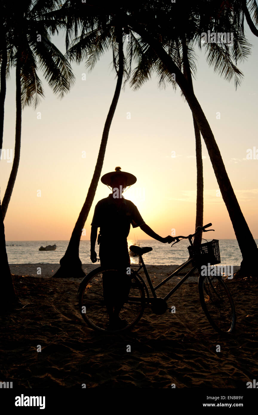Silhouette d'un homme portant un birman hat standing traditionnel par son vélo parmi les palmiers sur la plage de Ngapali Banque D'Images