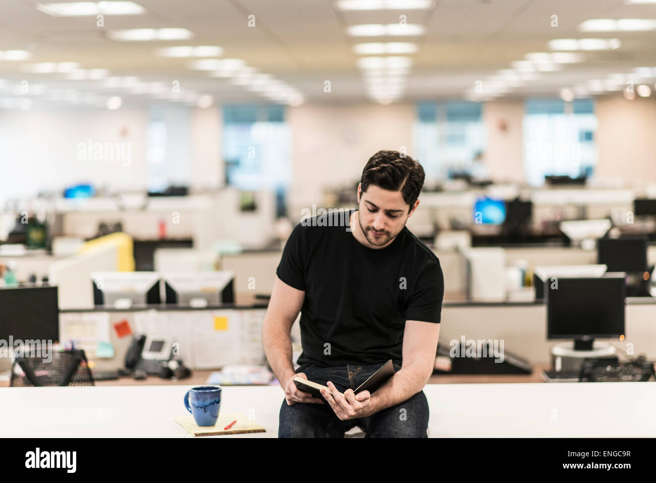 Un homme assis sur un bureau dans un bureau à la recherche d'un livre ou d'un journal. Banque D'Images