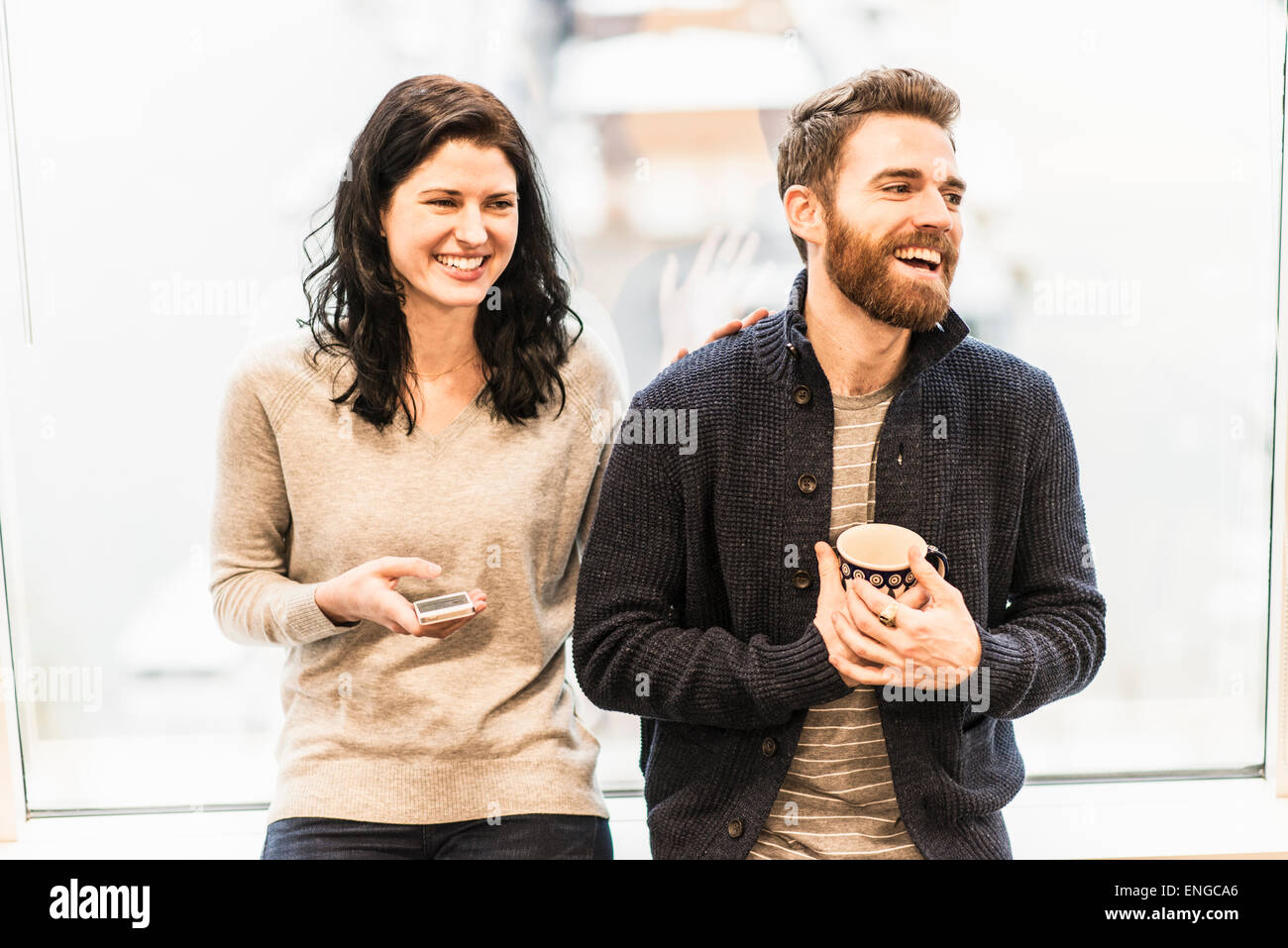 Une femme d'affaires par une fenêtre assis tenant un téléphone intelligent, le fait de parler à un homme tenant une tasse de café. Banque D'Images Une femme d'affaires par une fenêtre assis tenant un téléphone intelligent, le fait de parler à un homme tenant une tasse de café. Banque D'Images