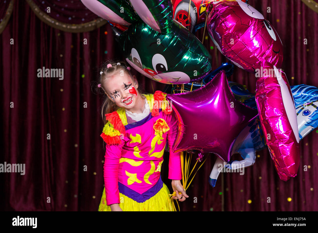 Smiling Blonde Girl costume coloré et composent Holding Bunch of aluminium ballons debout sur scène avec rideau rouge Banque D'Images