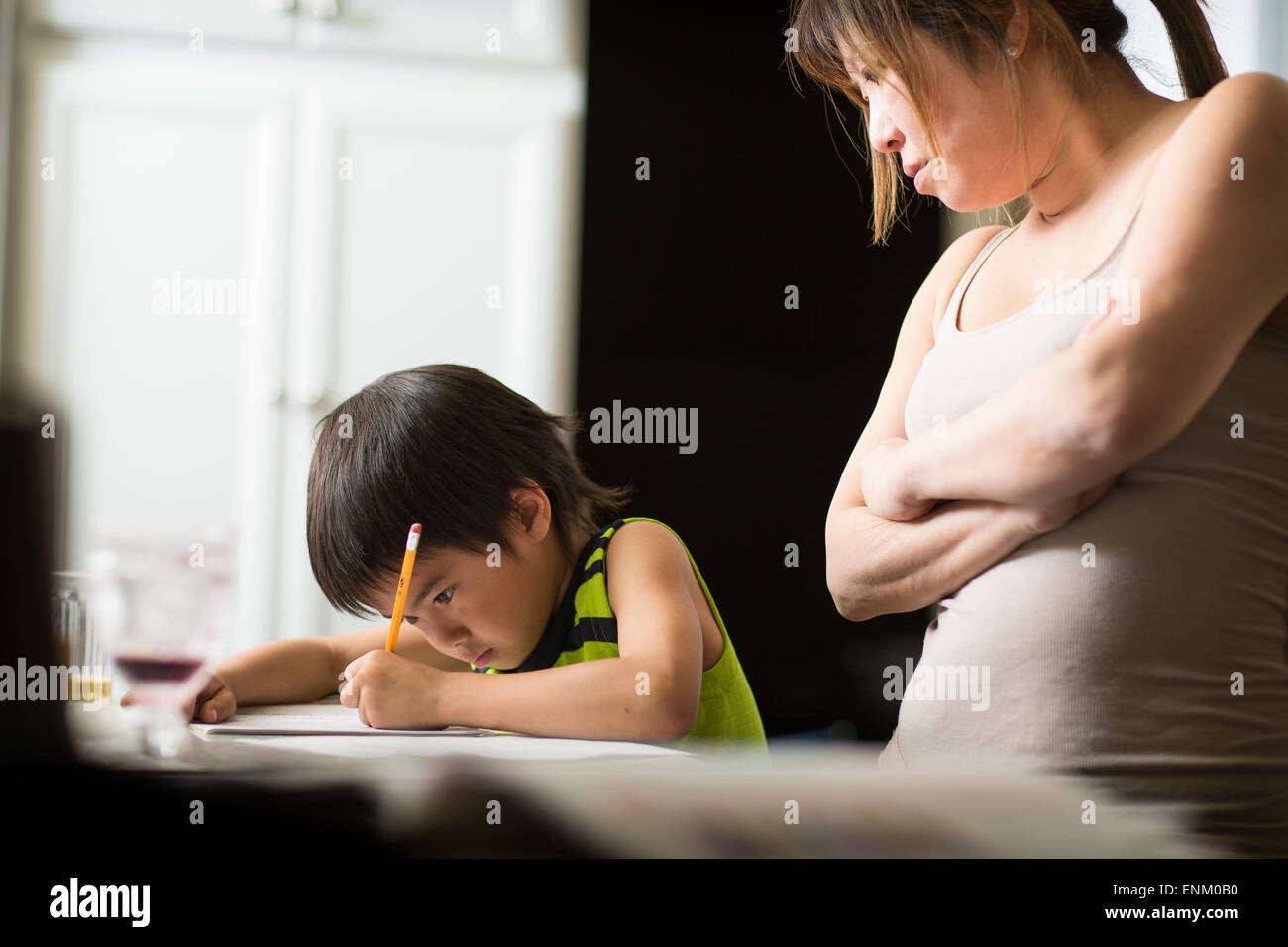 Un garçon Japonais Japonais études devoirs dans une cuisine comme sa maman regarde. Banque D'Images