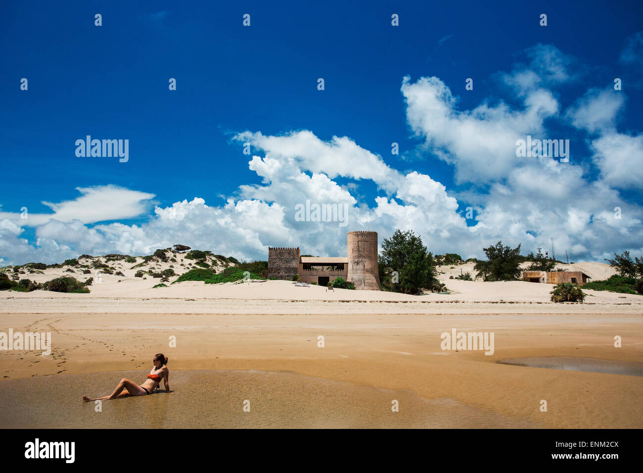 Océan Indien, Lamu, Kenya, Afrique. Une jeune femme en bikini se trouve en eau peu profonde sur une plage avec un vieux bâtiment de style château à distance. Banque D'Images