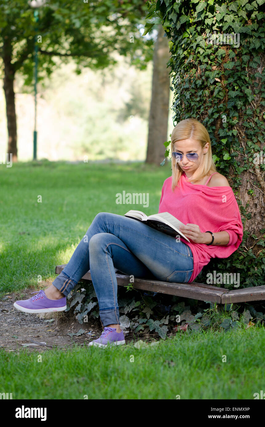 Jeune femme en bleu jeans avec des lunettes de soleil assis sur un banc et reading book Banque D'Images