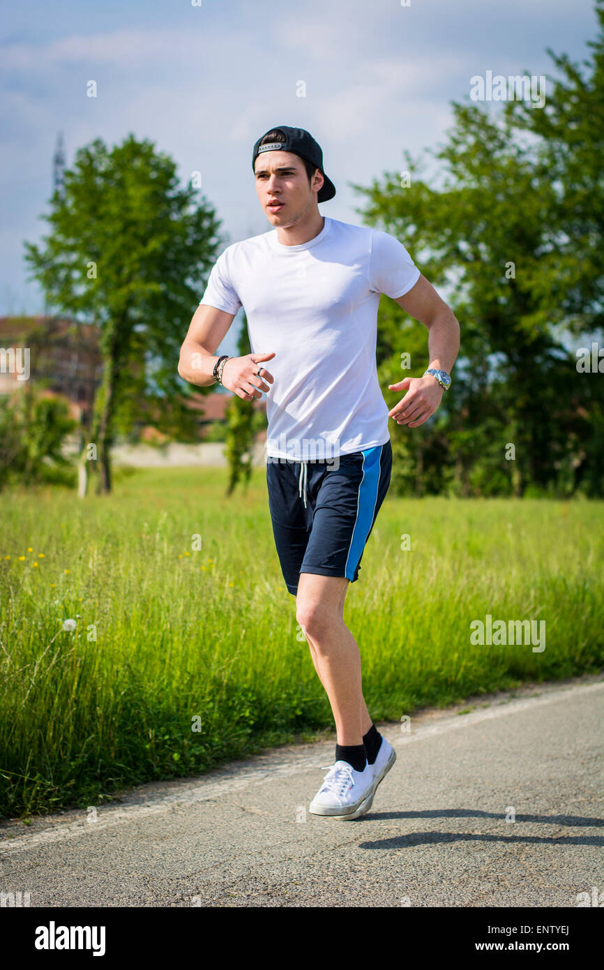 Beau jeune homme en marche et de la course sur route dans le pays en une journée ensoleillée, portant chemise blanche et chapeau de base-ball Banque D'Images