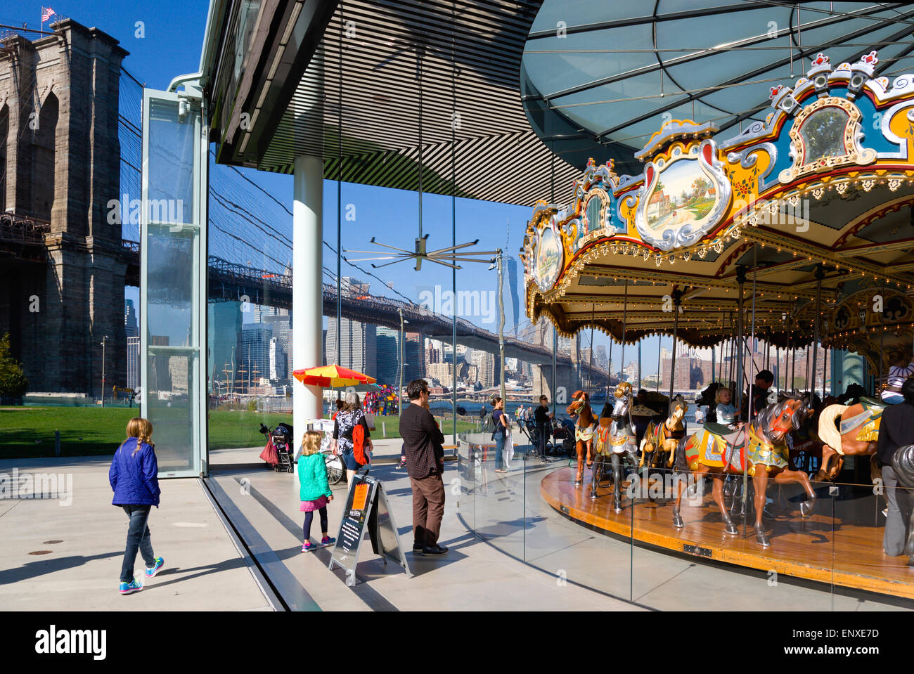 USA, New York State, New York, NYC, Brooklyn Bridge Park, les enfants et les adultes à Jane's Carousel dans un pavillon de verre dans l'Empire Fulton Ferry l'article ci-dessous le pont suspendu sur l'East River avec le Lower Manhattan skyline gratte-ciel au-delà. Banque D'Images