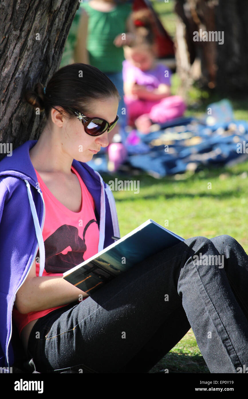 Une jeune femme assise contre un arbre en lisant un livre dans le parc. Banque D'Images