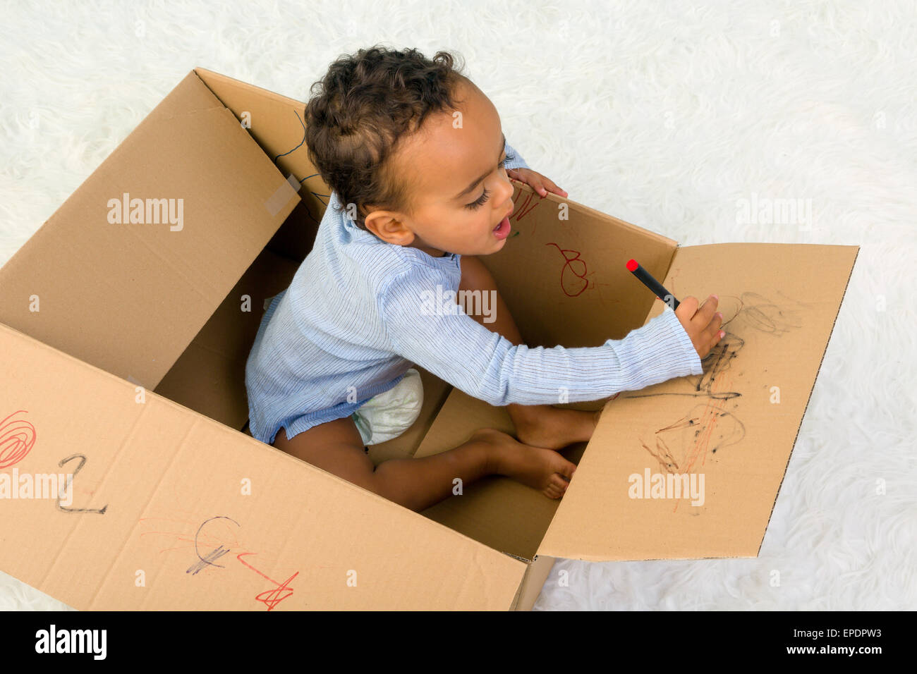 Mixed Race petit bébé garçon assis dans une boîte en carton à jouer avec des crayons Banque D'Images