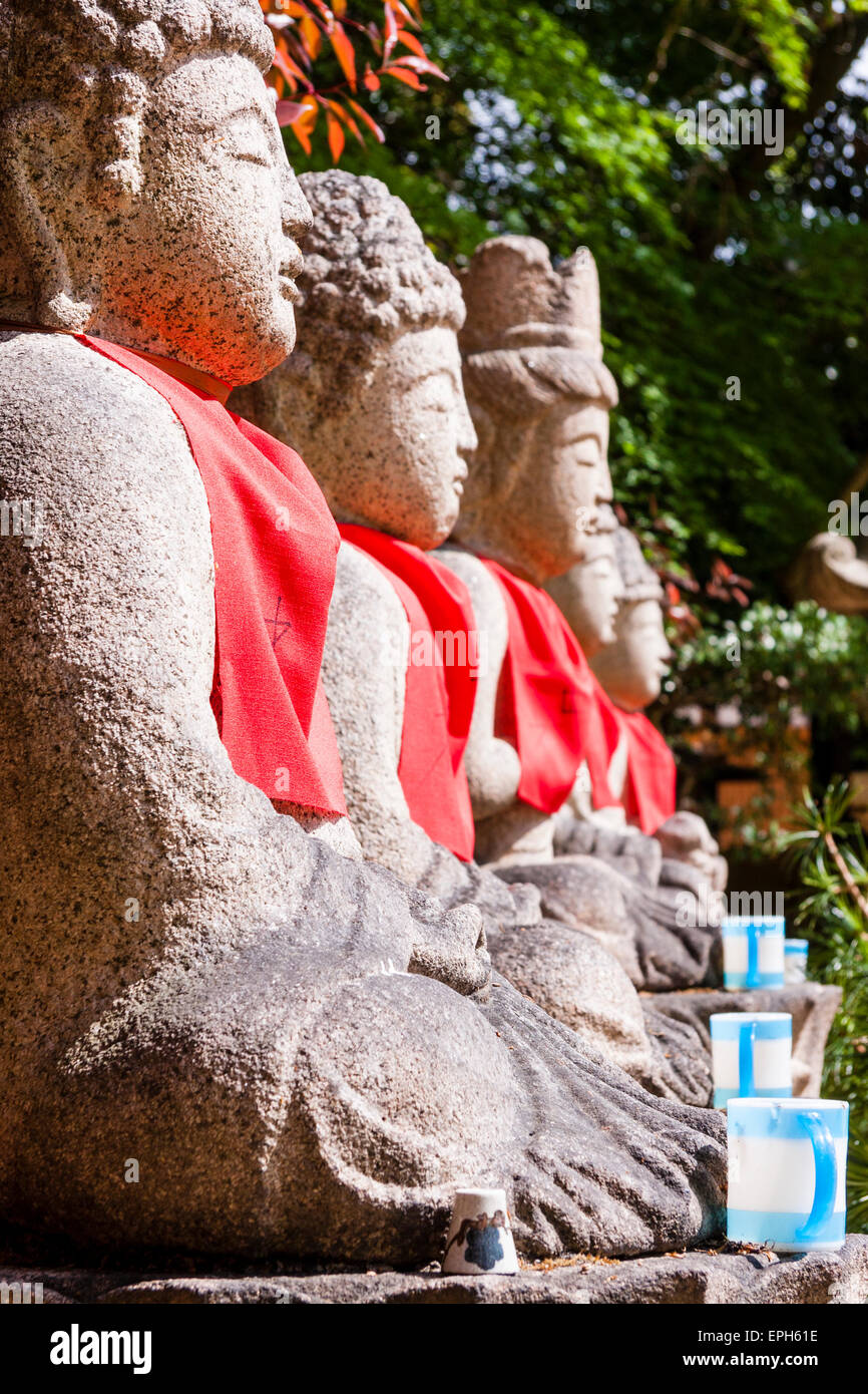 Vue rapprochée le long de quatre statues assises de Jozo bosatsu avec des bavoirs rouges en lumière, à l'extérieur au temple de Suma Dera à Suma, Japon. Banque D'Images