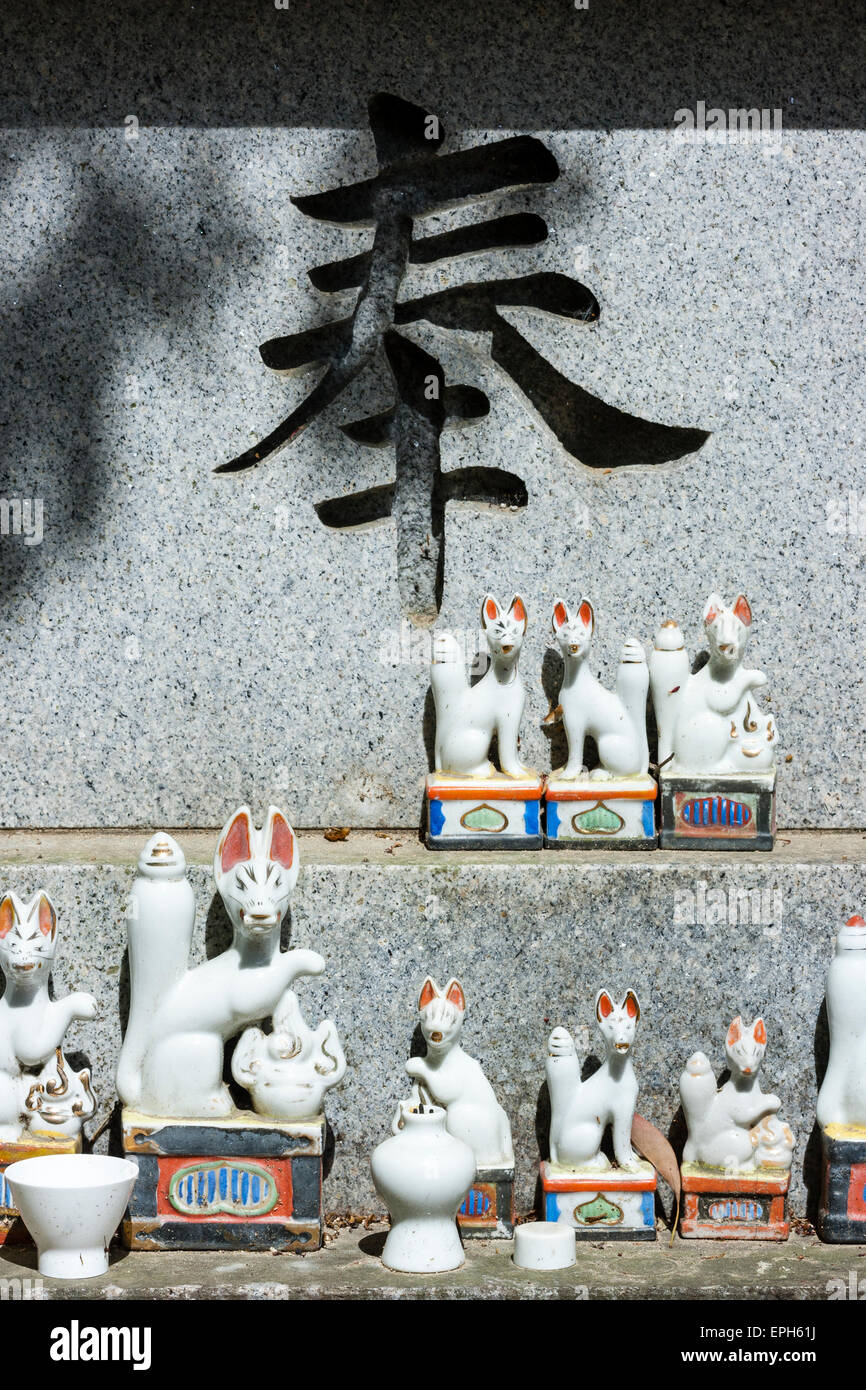 Petites statues de renard en porcelaine blanche et rouge, Inari, disposées sous un grand caractère kanji noir sur un bloc de marbre poli gris au temple Suma-dera, Japon. Banque D'Images