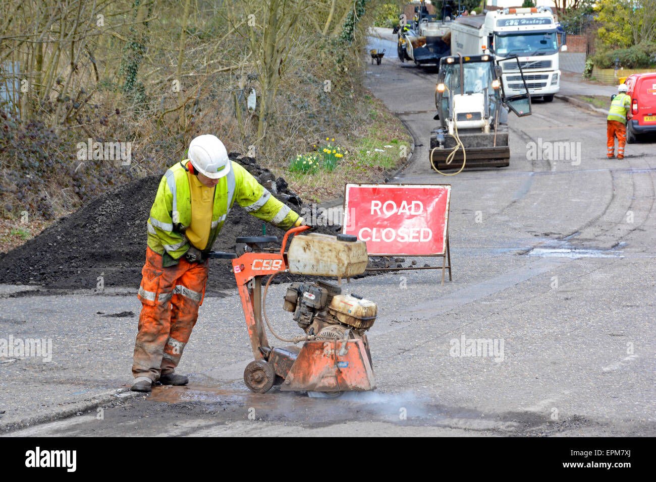 Village Road signe fermé et travailleur portant une machine de coupe de disque d'entraînement à haute visibilité faisant des travaux routiers réparation de nid-de-poule avant de resurfaçer Essex Angleterre Royaume-Uni Banque D'Images