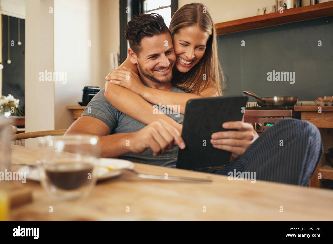 Couple smiling comme ils lisent une tablette ordinateur ensemble dans les matins dans la cuisine. Jeune homme et femme de rattrapage sur medi social Banque D'Images
