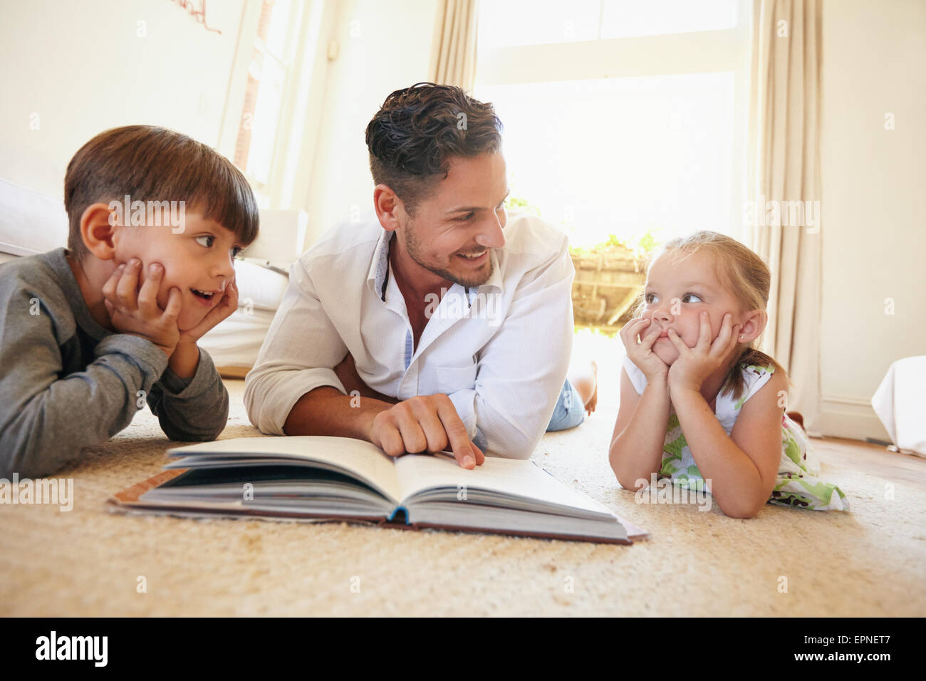 Tourné à l'intérieur du jeune homme avec deux enfants lisant un livre d'histoires. Famille étendue sur le sol avec un livre dans la salle de séjour. Banque D'Images