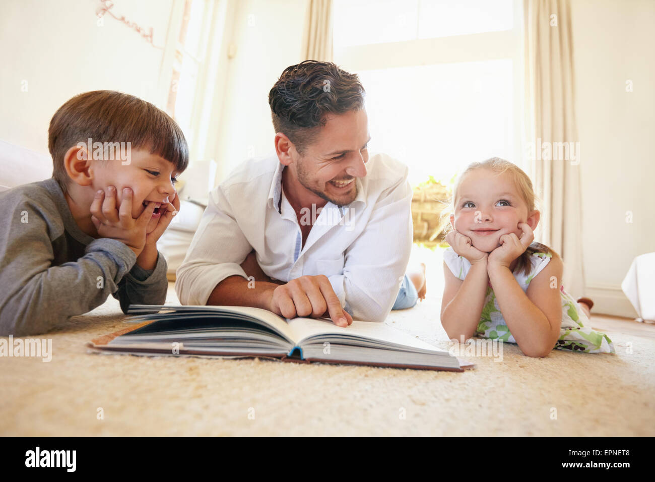 Portrait of happy young famille de trois couché sur étage avec un livre. Père de deux enfants lisant un livre d'histoires dans la salle de séjour à Banque D'Images