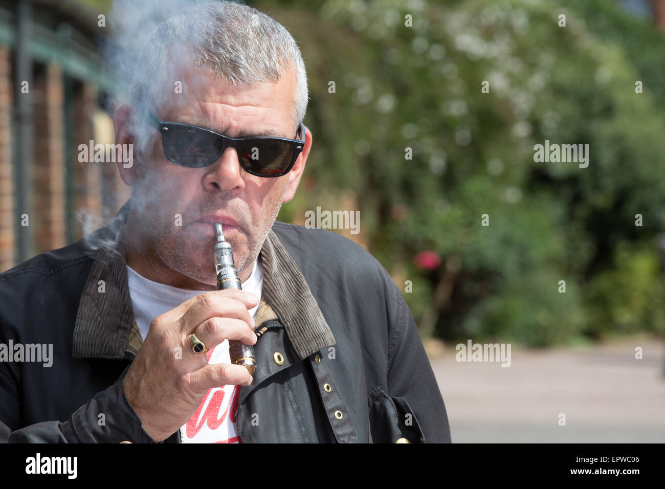 Homme d'âge moyen dans les lunettes de souffler sur un e-cigarette comme il se tient à l'extérieur dans l'avant-cour d'un bâtiment avec des arbres derrière h Banque D'Images