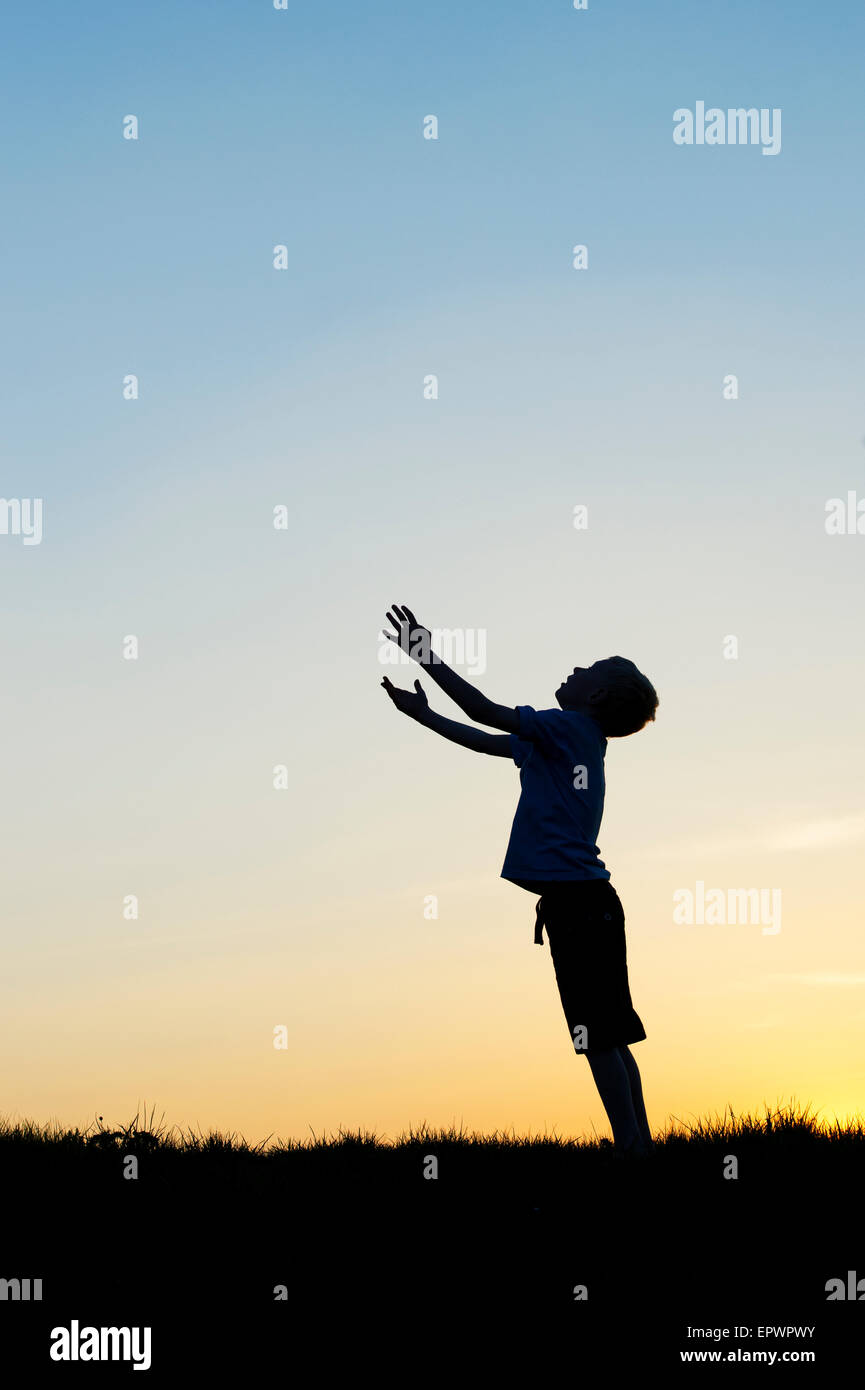 Boy looking up avec ses mains en l'air au coucher du soleil. Silhouette Banque D'Images