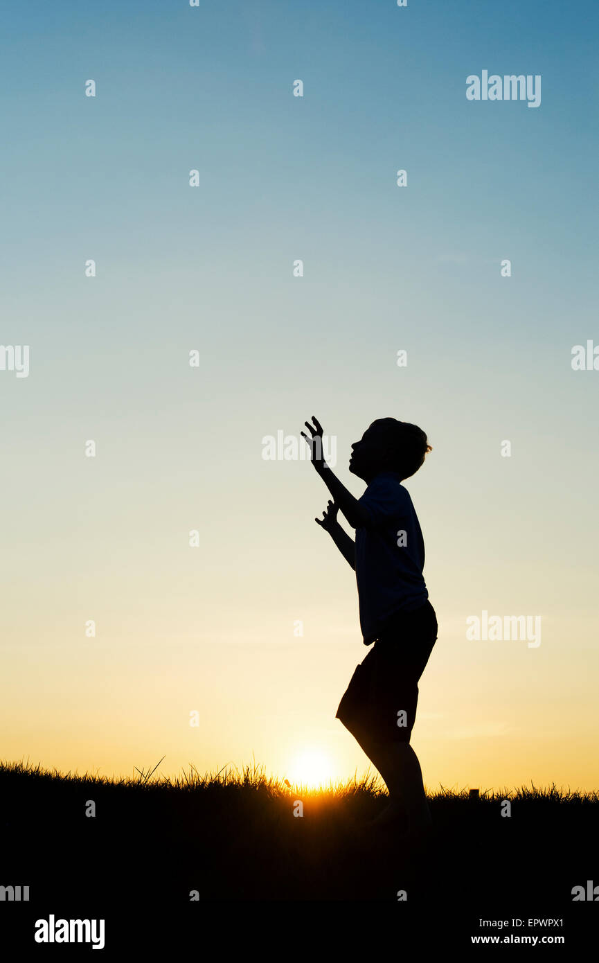 Boy looking up avec ses mains en l'air au coucher du soleil. Silhouette Banque D'Images