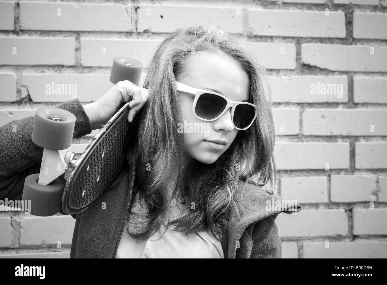 Adolescente blonde en jeans et lunettes détient plus de skateboard urban mur de briques, photo monochrome Banque D'Images