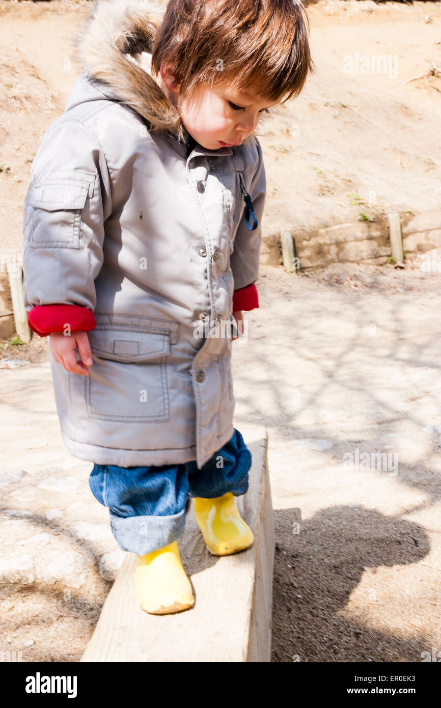 Enfant, garçon, 3-4 ans, extérieur par temps froid, portant un manteau  épais, avec des bateaux en caoutchouc jaune, tout en se tenant sur un banc  en bois, en regardant vers le bas